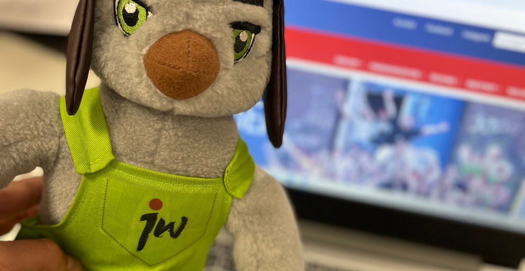 Unser Maskottchen Juwe guckt in die Kamera. Im Hintergrund ist ein PC zu sehen, auf dem unsere Website geöffnet ist. Vorne im Bild steht: Wir suchen dich!